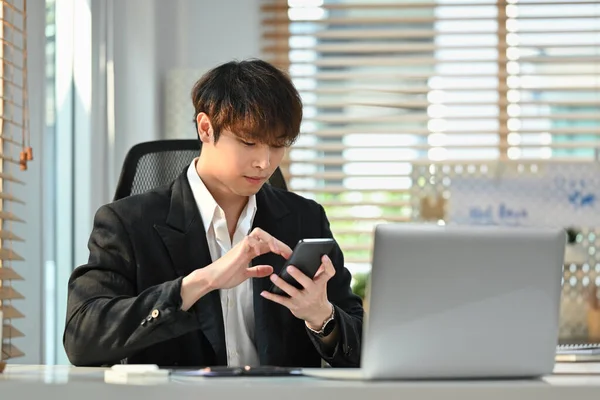 Millennial male corporate CEO in smart suit sitting in front of laptop at his office and using smart phone.