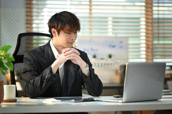 Attractive asian man corporate CEO in elegant suit watching online presentation on computer screen at workstation.