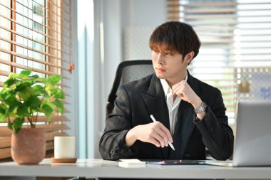 Successful young businessman sitting at workplace and looking through window, pondering idea or developing business strategy.