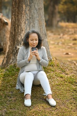 Satisfied middle aged woman holding paper cup of coffee resting on under tree. Authentic senior retired life, leisure activity concept.