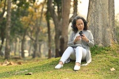 Pleasant middle age woman drinking coffee, relaxing under a tree on beautiful garden background.