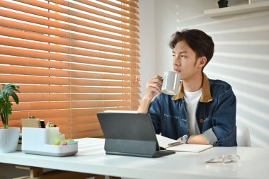 Thoughtful young asian man drinking coffee and looking through window, pondering idea in home office.