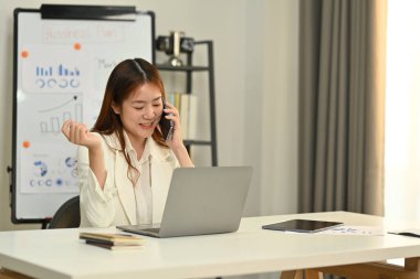 Güzel bir kadın finans danışmanı müşteriyle telefon görüşmesi yapıyor ve parlak ofiste dizüstü bilgisayar kullanıyor..