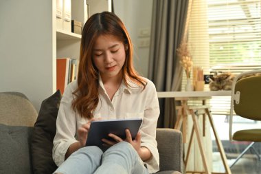 Günlük giysiler içinde güzel genç bir kadın dijital tablette kablosuz internete bakmaktan ve evdeki kanepede dinlenmekten zevk alır..