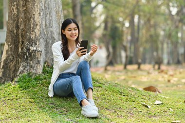 Ağacın altında cep telefonu kullanan güzel bir genç kadın. Ağaçların yaprakları manzaralı ve arka planda güneş ışığı var..