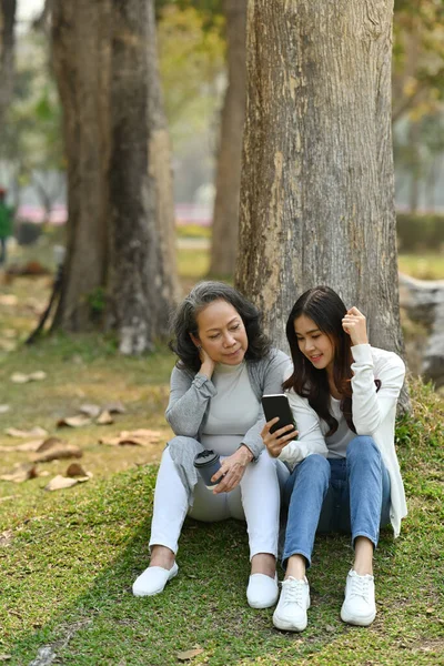 Mutlu kıdemli anne ve yetişkin kız akıllı telefon kullanarak yeşil çimenlerde otururken sakin doğa parkına karşı.