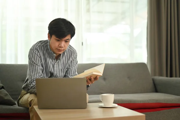 Relaxed asian man holding notebook and watching webinar or working online on laptop at home.