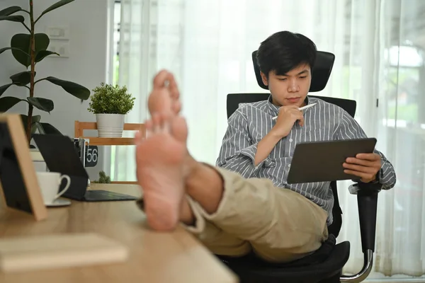 亚洲男性自由职业者穿着智能休闲装在现代家庭办公室使用数码平板电脑的形象 — 图库照片