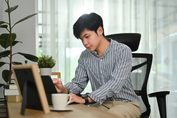Focused millennial business man using digital tablet browsing internet, work communicating online at home office.