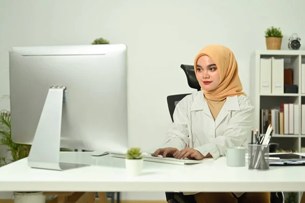 Smiling young muslim woman looking at computer monitor checking email, working with statistics at her office desk.