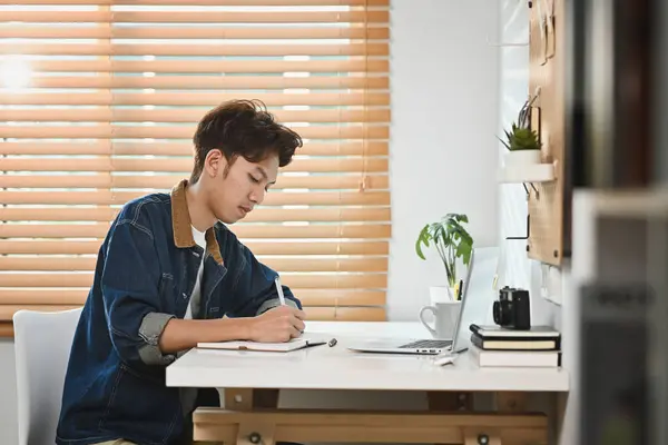 stock image Happy stylish man using laptop and making notes. Surfing internet, distance online study and remote work concept.