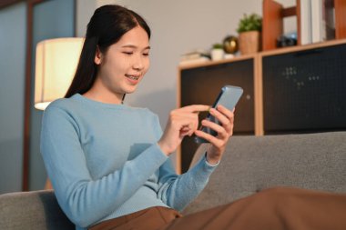 Sevimli genç bir kadın evdeki kanepede dinleniyor mesajlaşıyor, akıllı telefondaki kablosuz interneti tarıyor..