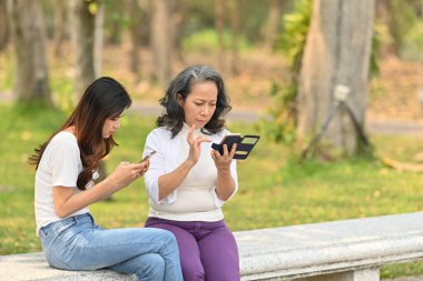 Sakin doğa parkında bir bankta otururken cep telefonu kullanan orta yaşlı kadın ve kızın görüntüsü.