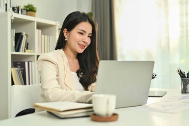 Modern ofiste masa başında oturan ve dizüstü bilgisayar kullanan başarılı, şık takım elbiseli genç iş kadını..