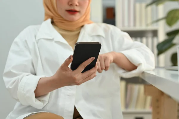 stock image Smiling young Muslim woman employee in hijab sitting at her working desk and chatting online on mobile phone.