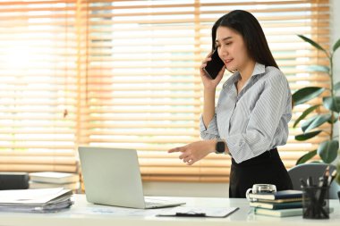 Güzel genç bayan finans danışmanı müşterisiyle telefonda konuşuyor ve bilgisayarını ofisinde kullanıyor..