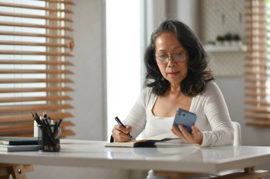 Cep telefonundan bilgi okuyan ve not defterine not alan orta yaşlı kadının portresi.