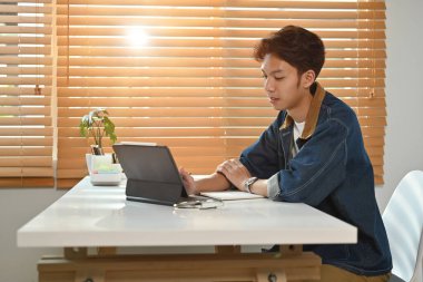 Evde dijital tablet kullanan kot ceketli genç Asyalı adam. Bağımsızlık, uzaklık, eğitim, e-öğrenme kavramı.