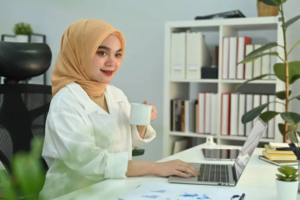 Muslim businesswoman using laptop computer, working with financial indicators at workplace.