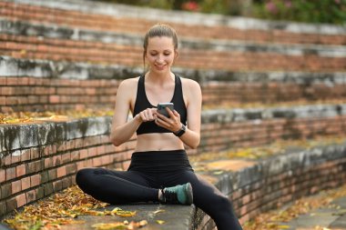 Gülümseyen atletik beyaz kadın akıllı telefon kullanıyor, dışarıda antremandan sonra dinleniyor. Fitness, spor ve sağlıklı yaşam tarzı kavramı.