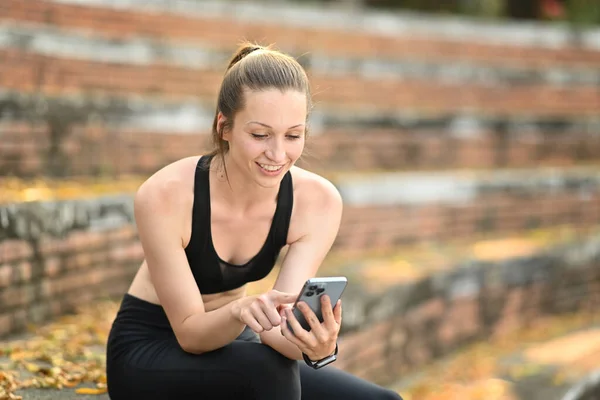 stock image Smiling young sportswoman using smart phone while relaxing on staircase outdoors. Fitness, sport and healthy lifestyle concept.