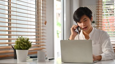 Yakışıklı genç işadamı, küçük işletme sahibi cep telefonuyla konuşuyor ve ofiste bilgisayarla çalışıyor..