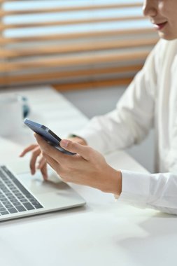 Gülümseyen bir iş adamının akıllı telefon kullanarak ofis masasında dizüstü bilgisayarla çalışırken görüntüsü..