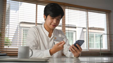 Yakışıklı yetişkin Asyalı tüketici kredi kartı ve akıllı telefon tutuyor, online alışveriş yapıyor veya online bankacılık işlemlerini yapıyor.