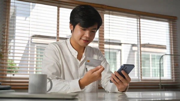 Handsome adult asian man consumer holding credit card and smartphone, shopping online or doing online banking transaction.