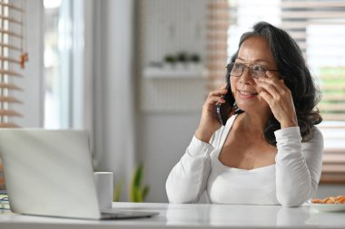 Başarılı üst düzey iş kadını, günlük kıyafetlerle evde çalışırken akıllı telefondan konuşuyor..