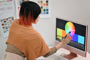 Yaratıcı ofisteki bilgisayar ekranında Asyalı kadın grafik tasarımcısının renk örnekleri arasından renk seçerken yan görünümü.