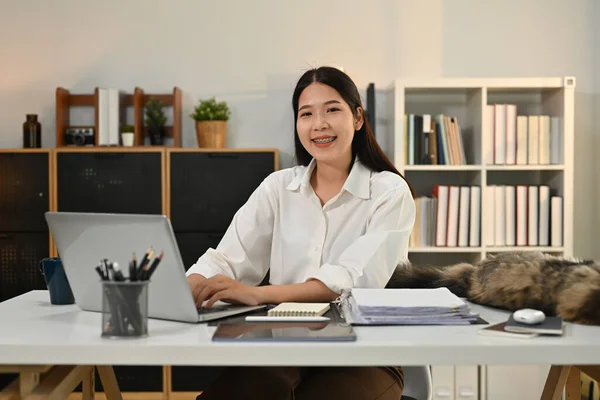 Portrait of asian female small business entrepreneur sitting at home office desk with laptop and smiling to camera.