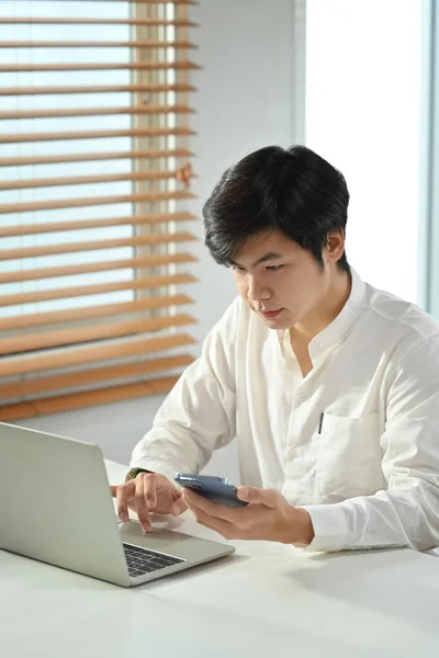 穿着白衬衫 手持手机 在办公桌前使用笔记本电脑的年轻英俊商人的画像 — 图库照片