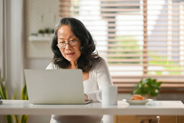 Beautiful senior woman entrepreneur working remote from home, reading email on laptop computer.