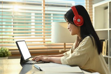 Gülümseyen Asyalı öğrenci kız kulaklık takıyor dijital tablette online ders seyrediyor. E-öğrenme ve teknoloji kavramı.