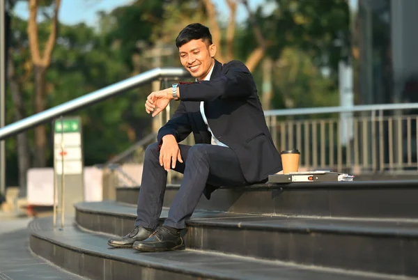 Full length of young businessman sitting on stairs outside office building and checking time on his wristwatch.