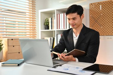 Milenyum Asyalı erkek girişimci laptop ekranına bakıyor ve günlük planlamacıya kayıt tutuyor..