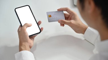 Kredi kartı ve akıllı telefonu olan, online alışveriş yapan ya da online banka işlemleri yapan bir adamın omuz üstü görüntüsü.