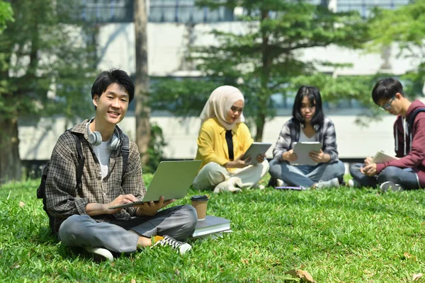 年轻的亚裔学生在大学大楼前的绿草上使用笔记本电脑 技术和生活方式概念 — 图库照片