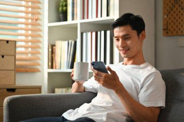Rahat Asyalı bir adam evde kanepede otururken kahve içiyor ve sosyal medyayı akıllı telefonuyla kontrol ediyor..