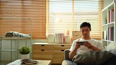 Dolly 'nin çektiği kaygısız Asyalı adam kanepede oturmuş akıllı telefondan video oyunu oynuyor. Teknoloji, eğlence ve eğlence aktivitesi.
