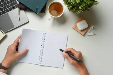 Çalışma masasındaki boş spiral not defterine yazı yazan kadın çalışanın yukarıdan görünüşü.