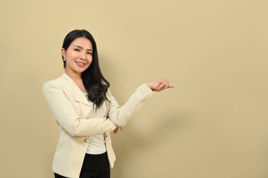 İş kıyafetleri içindeki güzel Asyalı kadın ürün veya metin için fotokopi alanını işaret ediyor, bej arka planda izole edilmiş..