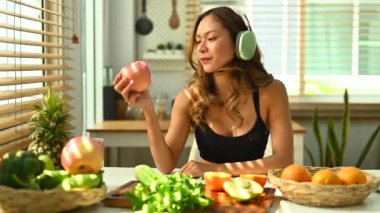 Spor kıyafetleri ve kulaklık takan genç kadın mutfak masasında sağlıklı yeşil elma yiyor..