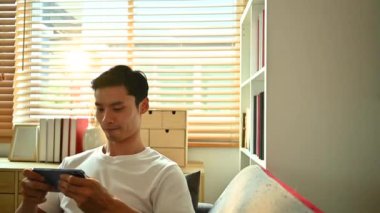 Kaygısız Asyalı adam kanepede oturmuş akıllı telefondan video oyunu oynuyor. Teknoloji, eğlence ve eğlence aktivitesi.
