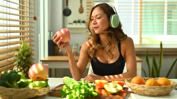 身穿运动服 头戴耳机的年轻健康女性在厨房桌上吃着健康的绿色苹果 — 图库视频影像