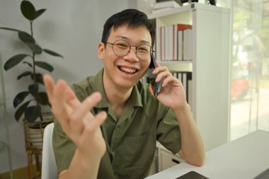 Gözlüklü, gülümseyen milenyum adamı cep telefonuyla konuşuyor, laptopuyla çalışma masasında oturuyor..