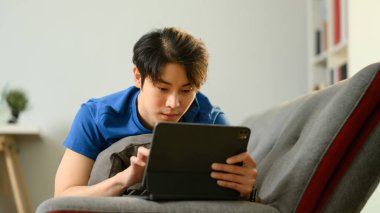 Dijital tablet kullanarak kanepede uzanan sıradan bir Asyalı. İnsanlar, teknoloji ve yaşam tarzı.