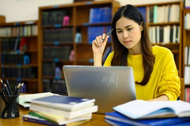 Masada oturan Asyalı kız öğrenci ders kitapları ve laptopla düşünceli arama çözümü, sınava hazırlanıyor..