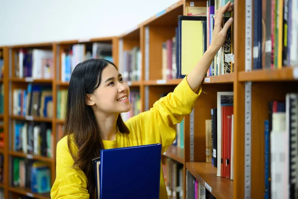 Asian female student picking up a book, choosing books from shelf in library. People, knowledge and education concept.